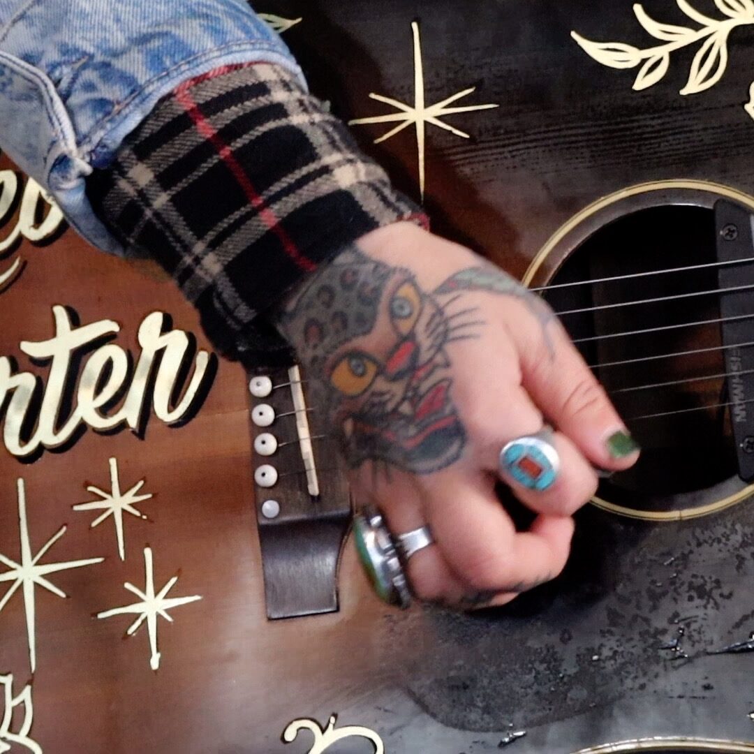 Close up photo of a woman's ringed hand, tattooed with a colorful big cat, strumming an ornately decorated vintage acoustic guitar including her name Rebecca Porter in fancy letters and space age stars, miked for recording.