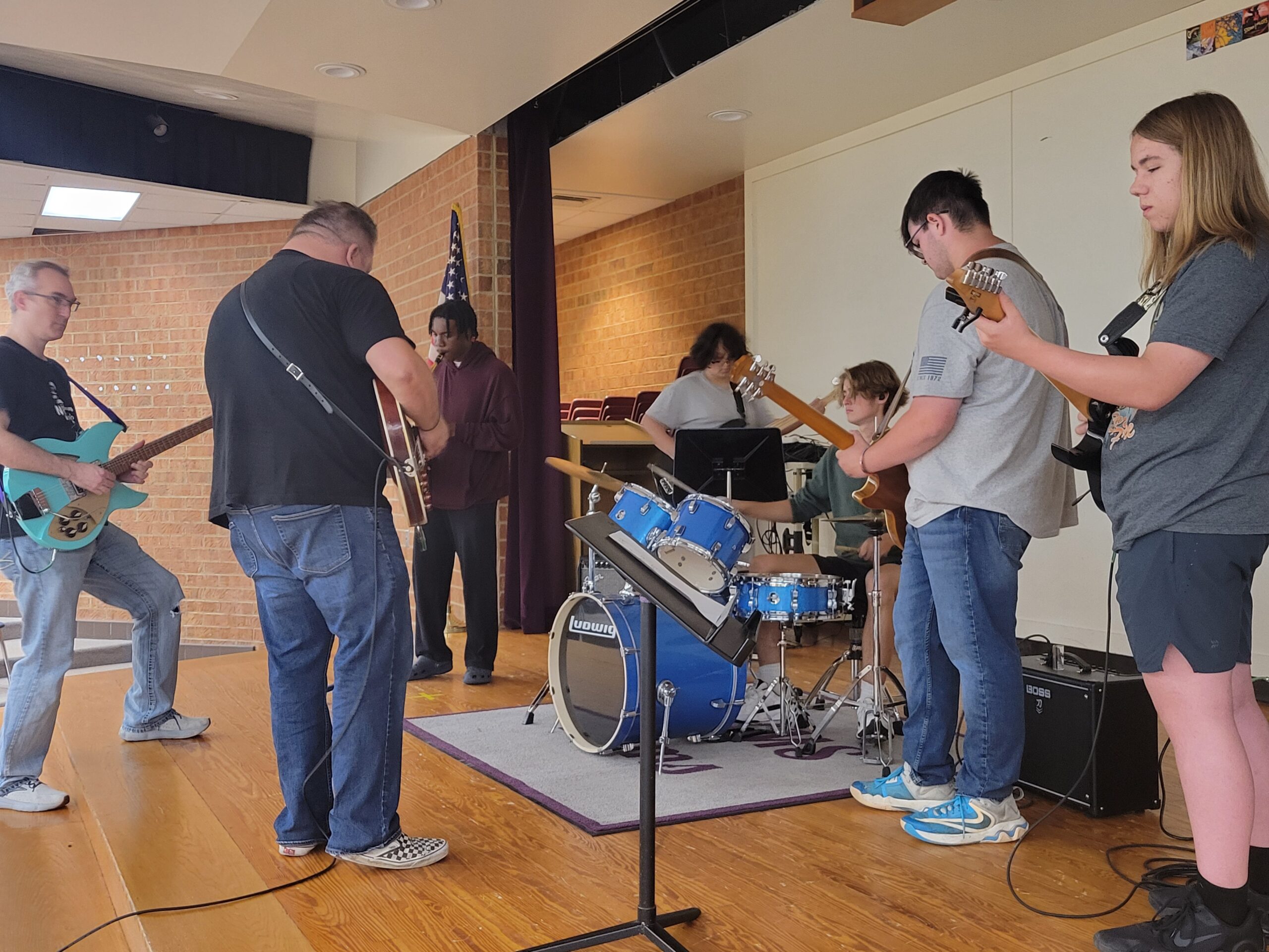 Two adult instructors play electric guitars along with high school rock band students on stage in a small indoor auditorium